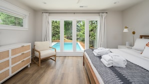 A bedroom with a bed, a chair, a bedside table, a lamp, and a view of a swimming pool through large windows. - Close to the Beach! (Hilton Head Island)