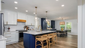 A modern kitchen with white and blue cabinets, a large island with bar stools, and pendant lighting. - Close to the Beach! (Hilton Head Island)