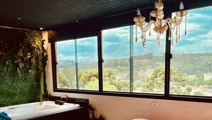A bedroom with a large bed, a view of the countryside through large windows, a chandelier, and a green wall with plants. - Maison France (Campos do Jordão)