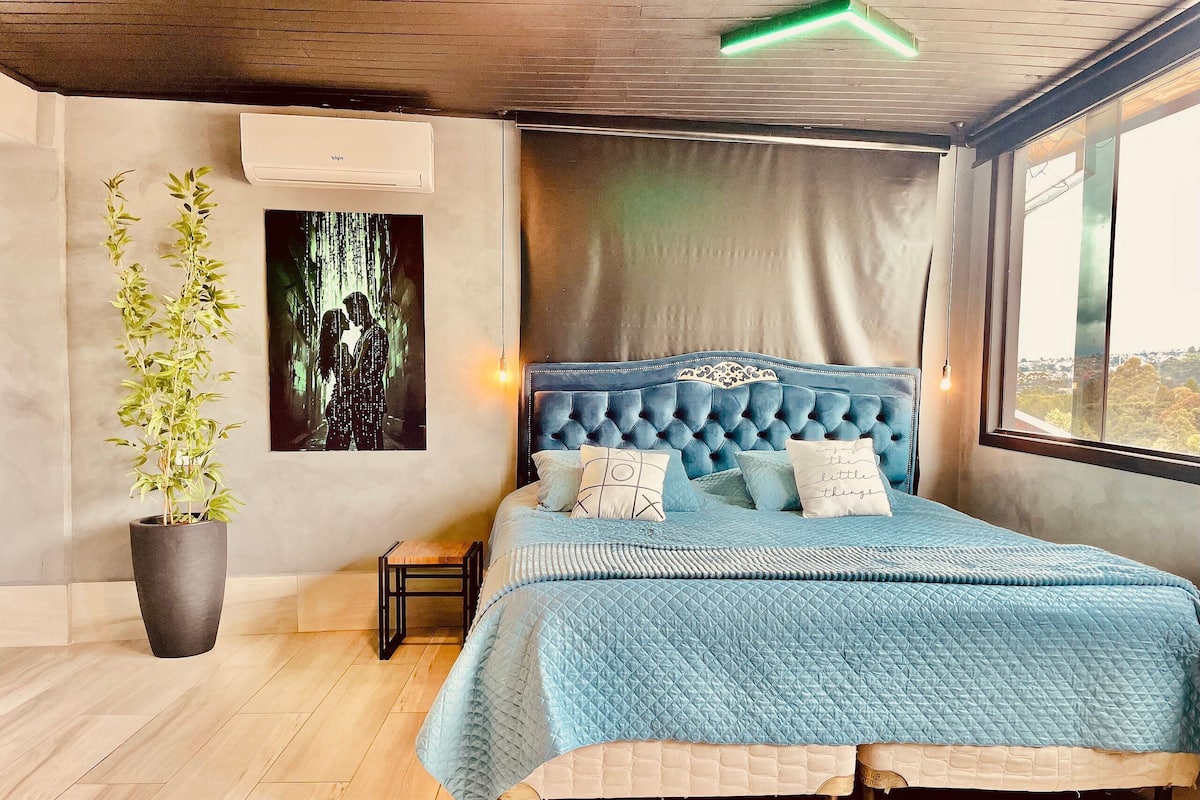 Une chambre avec un grand lit, une plante en pot, une petite table et un tableau accroché au mur.