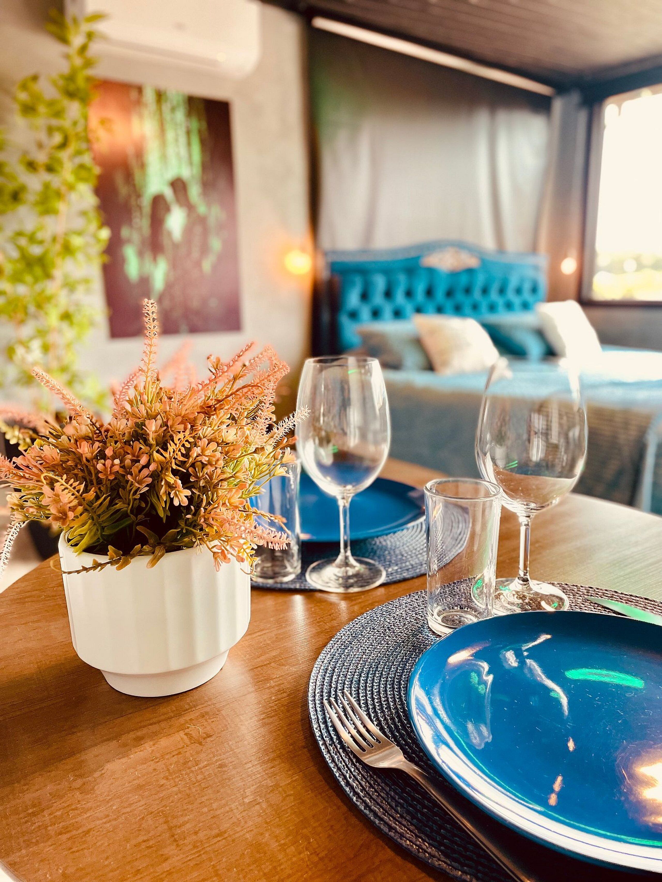 Une table à manger dressée avec un pot blanc contenant des fleurs séchées, deux verres à vin et des assiettes bleues.