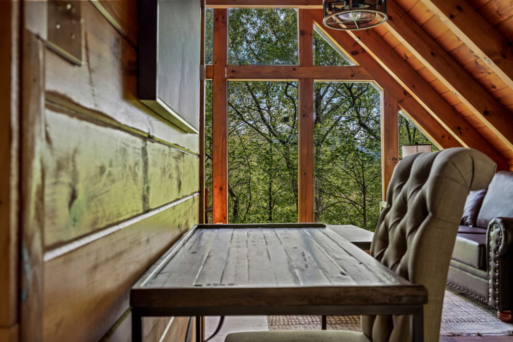 Interior de madera con una mesa y un banco, ventanas amplias que dan a un paisaje de árboles.