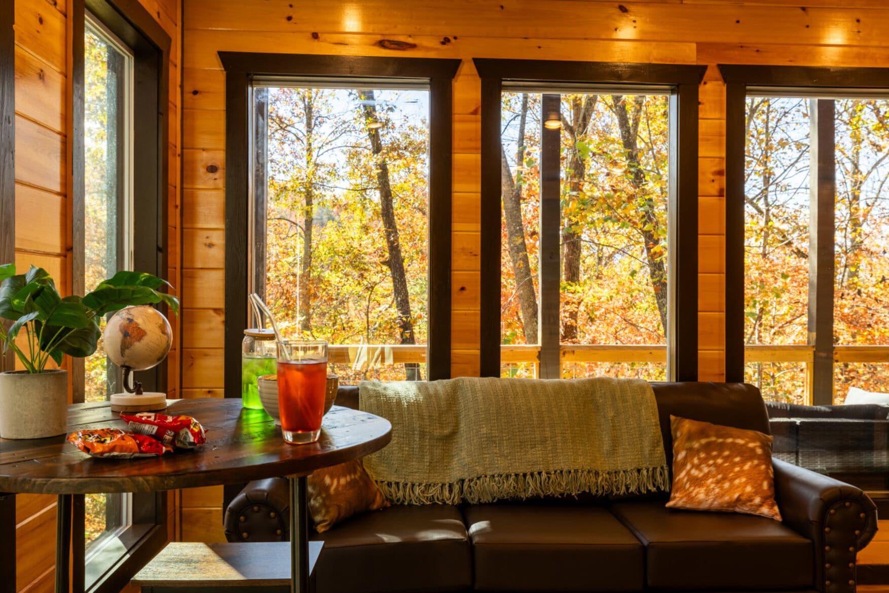 Interior acogedor de una cabaña con detalles de madera, una mesa redonda con un globo terráqueo y aperitivos, un sofá de cuero con una manta, y ventanas amplias con vistas a follaje otoñal.