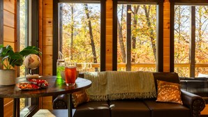 L’intérieur chaleureux d’un chalet, avec un décor en bois, une table ronde ornée d’un globe terrestre et de quelques collations, un canapé en cuir agrémenté d’un plaid, et de grandes fenêtres offrant une vue sur le feuillage automnal.