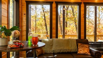 L’intérieur chaleureux d’un chalet, avec un décor en bois, une table ronde ornée d’un globe terrestre et de quelques collations, un canapé en cuir agrémenté d’un plaid, et de grandes fenêtres offrant une vue sur le feuillage automnal.