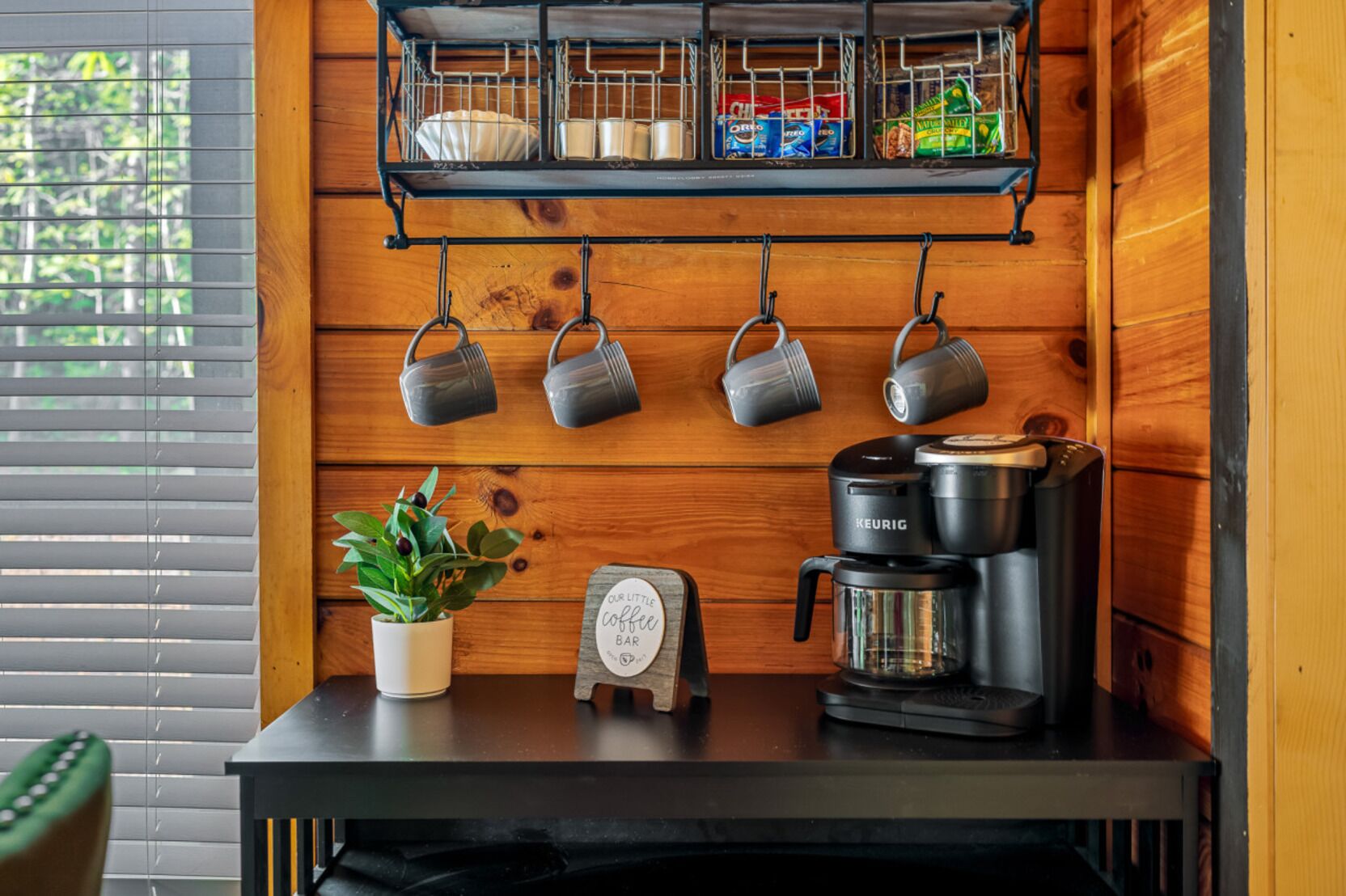 A kitchenette with a coffee maker, mugs, a potted plant, and a sign that reads "Our Little Coffee Bar".