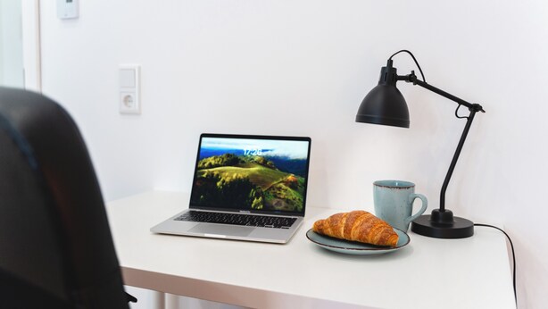 Un ordinateur portable posé sur un bureau, avec un fond d’écran représentant un paysage, une tasse bleue et un croissant.