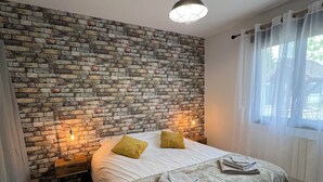 Een slaapkamer met een muur in baksteenmotief, een bed met een beige sprei, twee kussens, een nachtkastje met een lamp en een raam met dunne gordijnen.