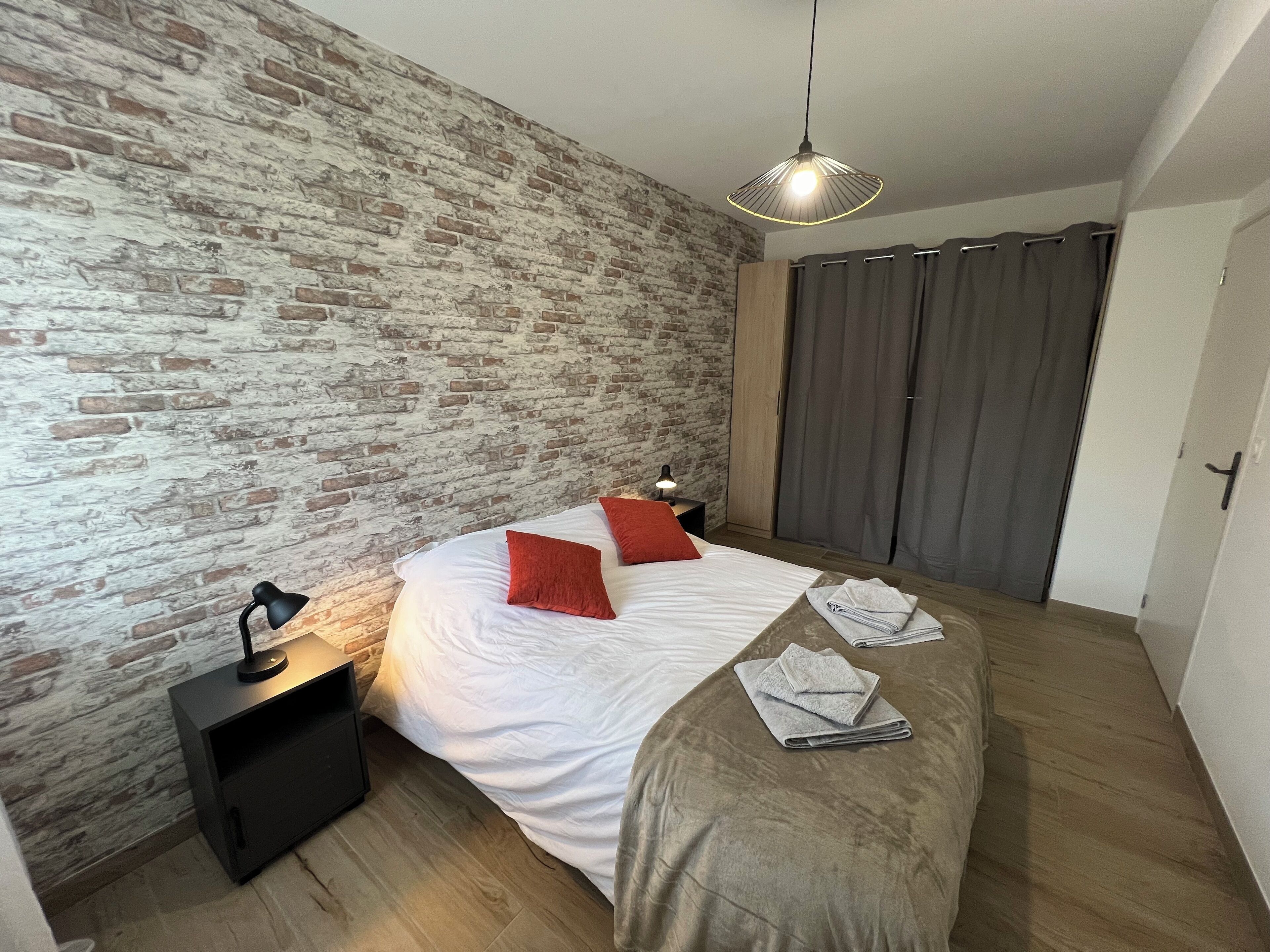 Une chambre avec un mur en briques, un lit avec une literie blanche et des oreillers rouges, une table de chevet avec une lampe et un luminaire suspendu.