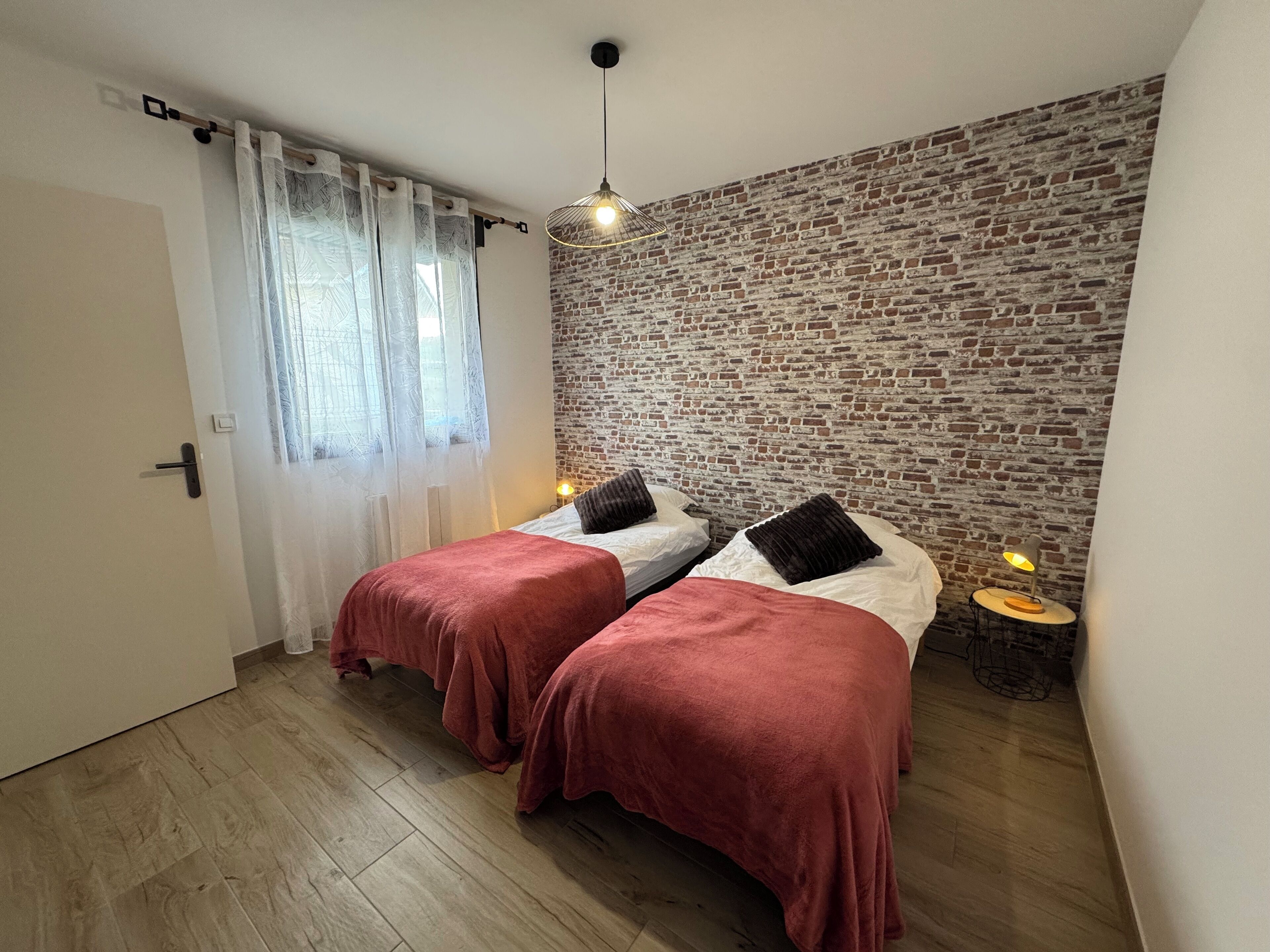 Une chambre avec deux lits, un mur en briques, un plancher en bois, une fenêtre avec des rideaux et un luminaire suspendu.