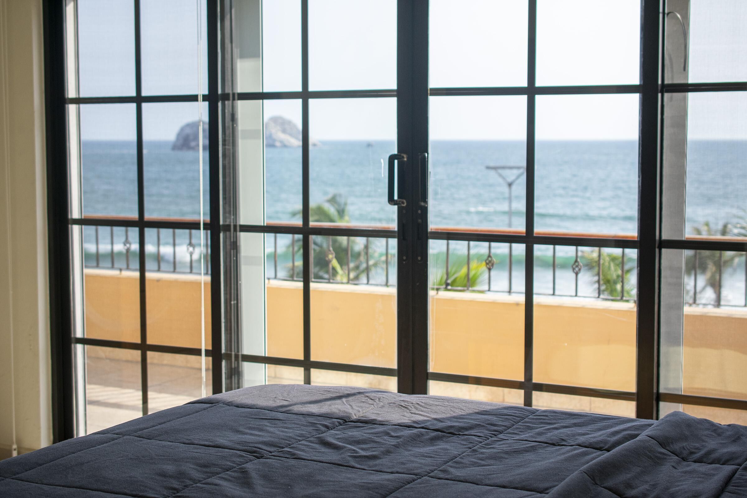 Een kamer met een bed, grote ramen met uitzicht op de oceaan en een balkon met een hekwerk.