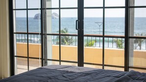 Ein Zimmer mit einem Bett, großen Fenstern mit Meerblick und einem Balkon mit Geländer.