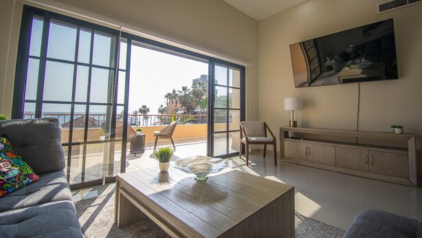 Ein geräumiges Wohnzimmer mit einer großen Glasschiebetür, die zu einem Balkon führt, einem an der Wand montierten Flachbildfernseher und einem Holztisch mit Dekorationen.