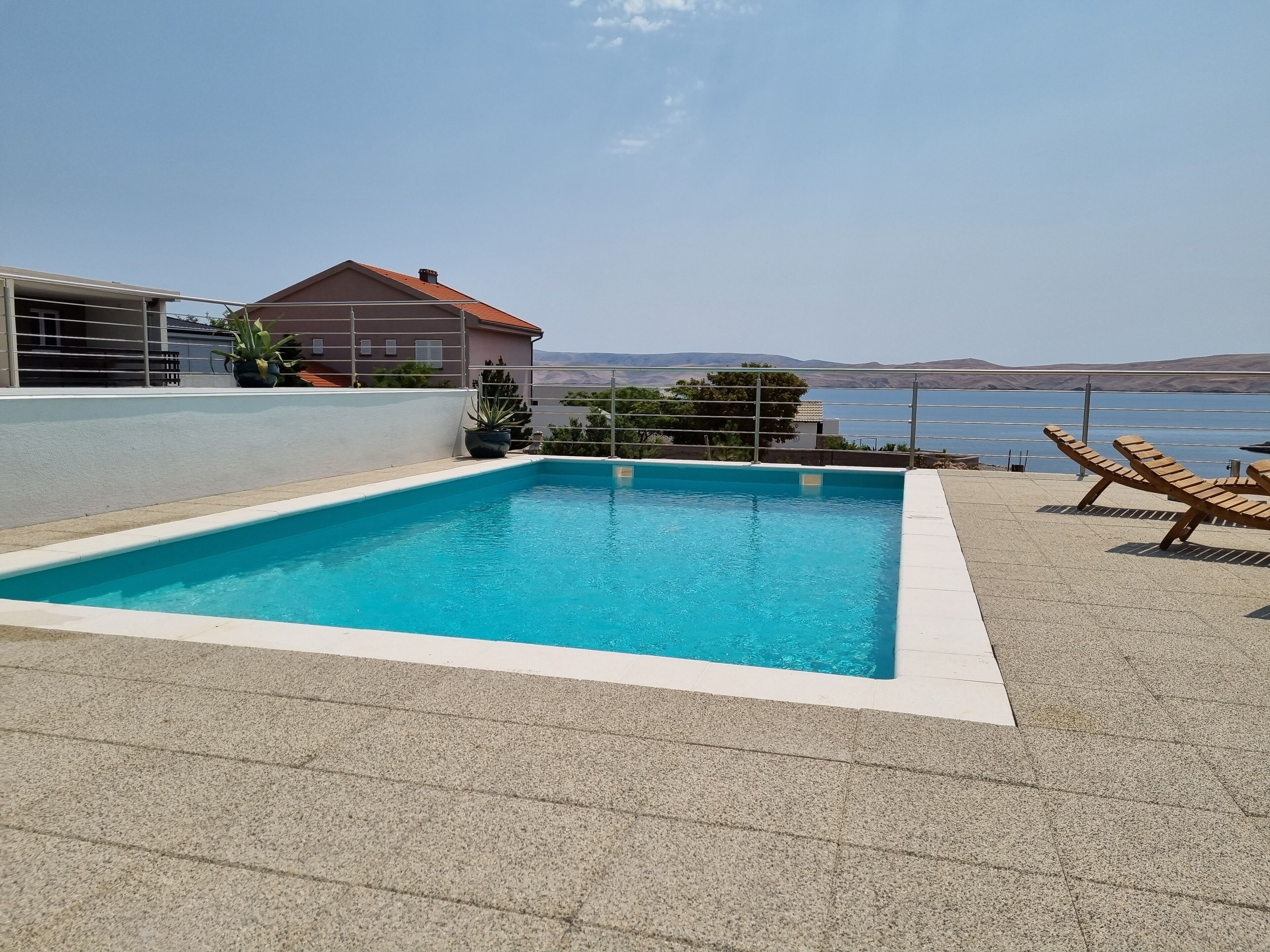 A rectangular swimming pool with a clear blue water, surrounded by a tiled deck. There are two wooden lounge chairs on the right side of the pool. In the background, there is a house with a red roof and a view of the sea.