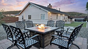 An outdoor patio with a fire pit table and chairs, a house in the background, and a clear sky.