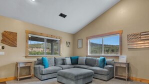 A living room with a sectional sofa, two windows with blinds, a ceiling vent, and a welcome sign.