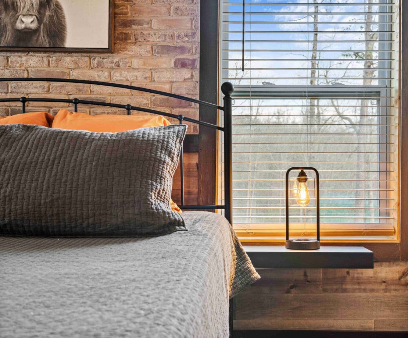 A bedroom with a brick wall, a bed with grey bedding, orange pillows, and a window with blinds and a hanging light fixture.