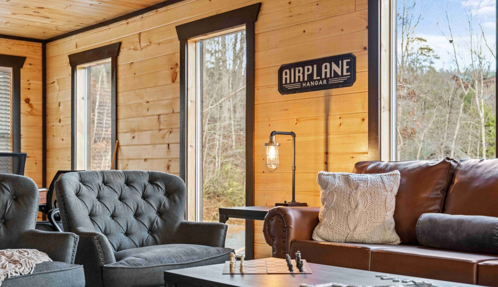 A cozy cabin interior with a large window, a sign reading "AIRPLANE HANGAR," and a leather sofa.