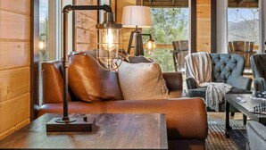 A cozy cabin interior with a wooden interior, a sofa with cushions, a table with a lamp, and a view through large windows. - Hidden Gem Masterpiece By Ghosal Luxury Lodging  (Gatlinburg)