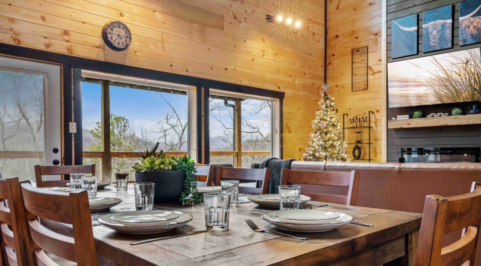 A wooden interior with a dining table set for four, large windows, a Christmas tree, and a clock on the wall.