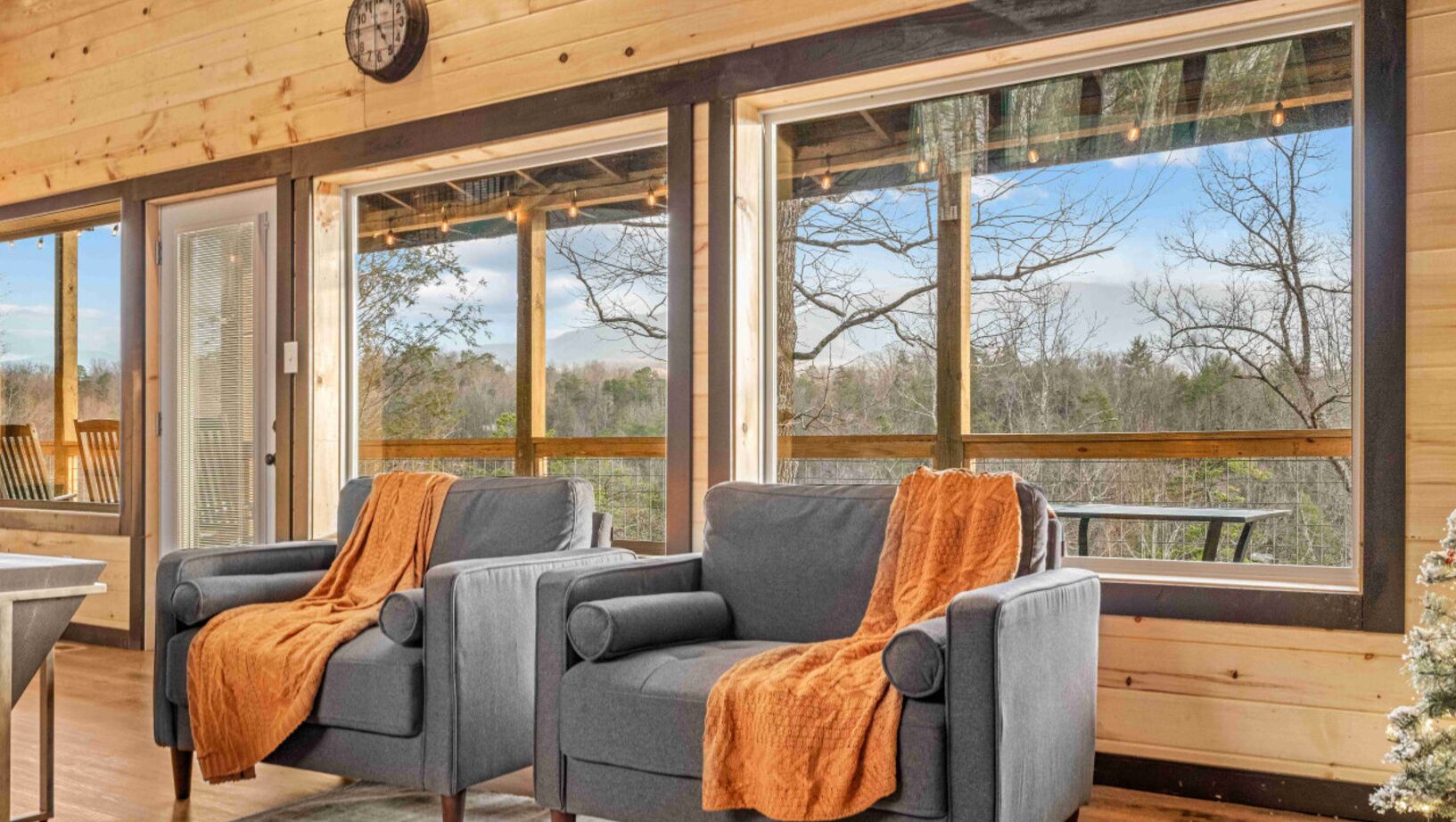 A cozy deck area with two gray sofas and a wooden table, large windows offering a view of trees and a clear sky.