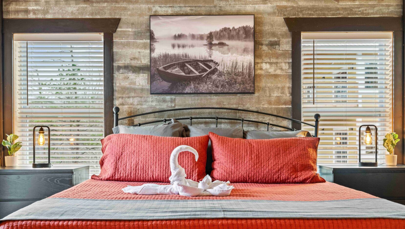 A bed with a red quilt and pillows, a swan-shaped pillow, and a framed picture of a boat on water.