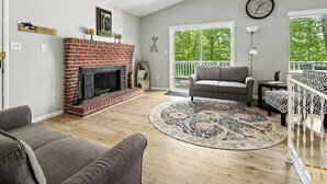 A living room with a fireplace, a sofa, a patterned rug, and a view of trees through large windows. - Charming waterfront retreat on Buggs Island/Kerr Lake—perfect lakeside living! (Clarksville)
