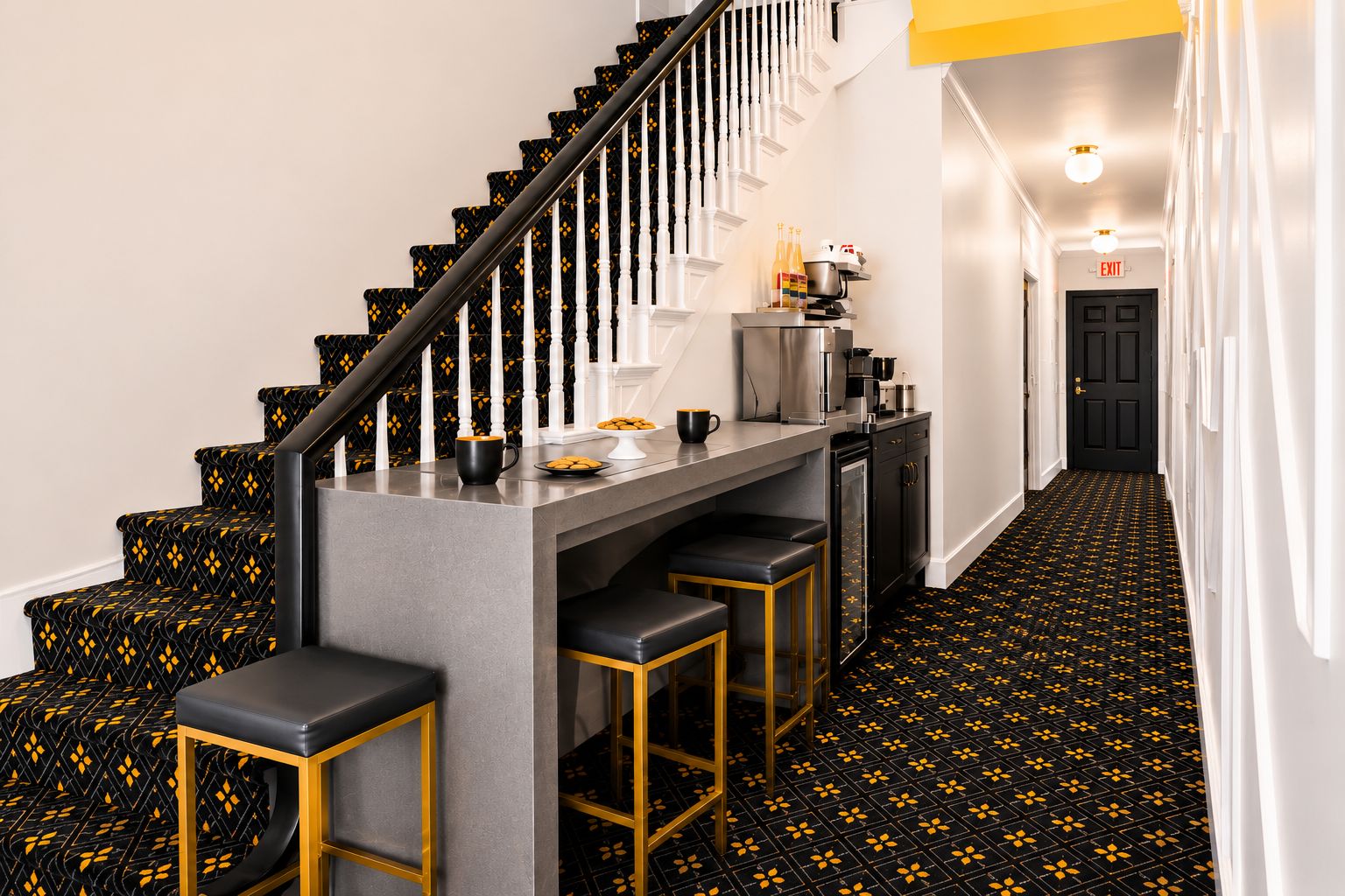 Una escalera con alfombra de estampado negro y amarillo, una zona de bar con taburetes y una cocina con electrodomésticos.