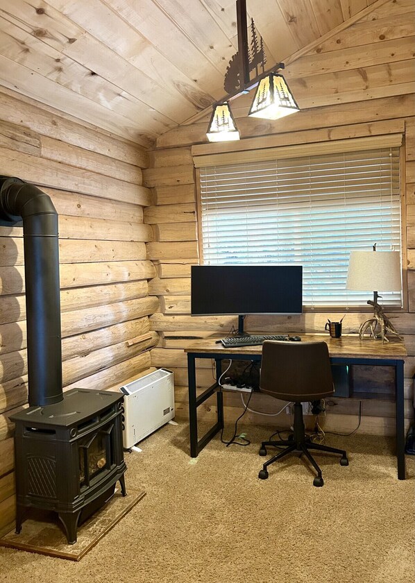 A log cabin interior with a wood-burning stove, a desk with a computer, and a window with blinds.