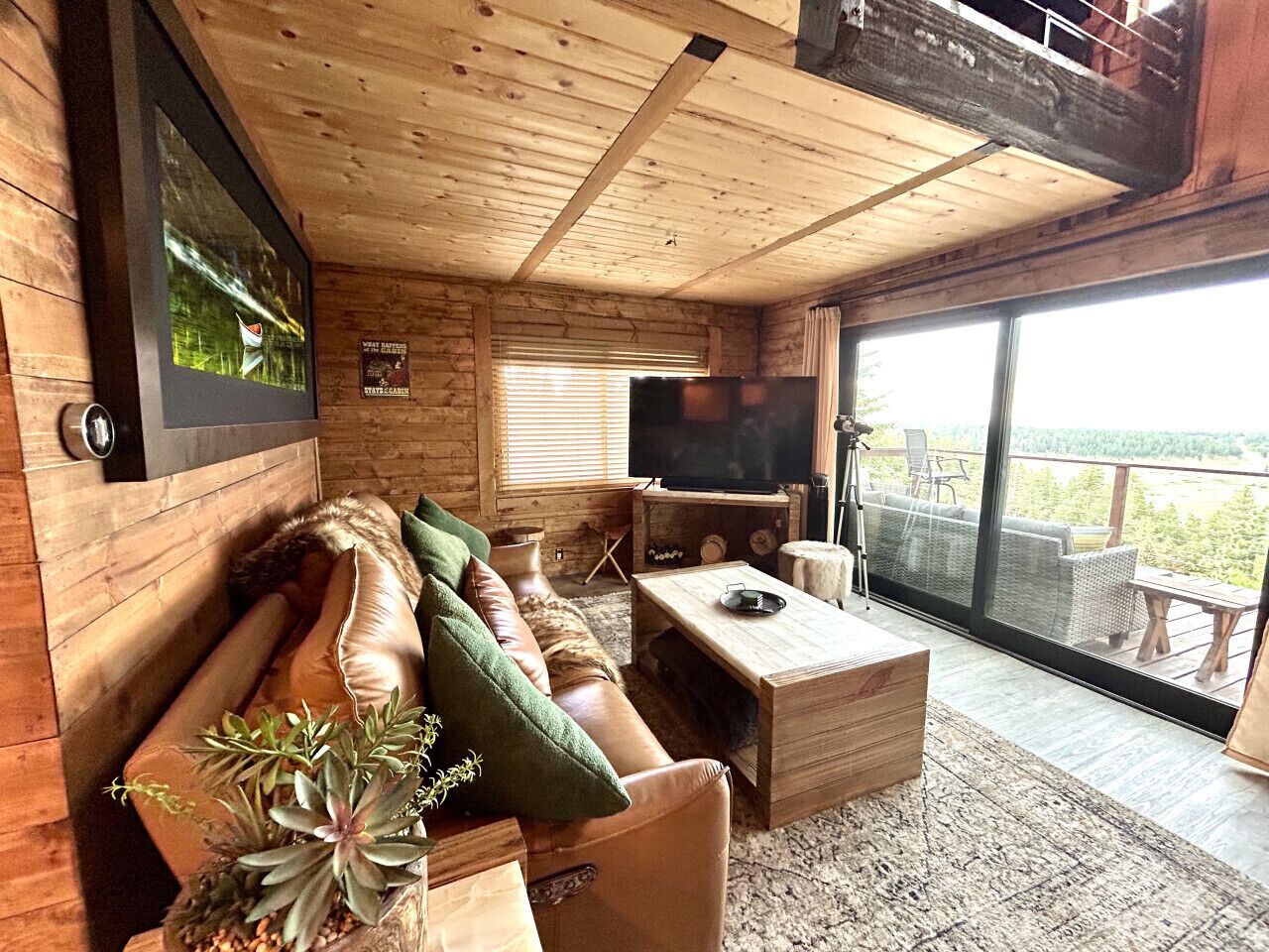 A cozy cabin interior with a wooden ceiling, a large TV, a sofa with cushions, and a sliding glass door leading to an outdoor deck.