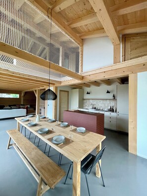 Een moderne keuken met een houten eiland, een eethoek met een lange tafel en banken, en een hoge plafond met houten balken.