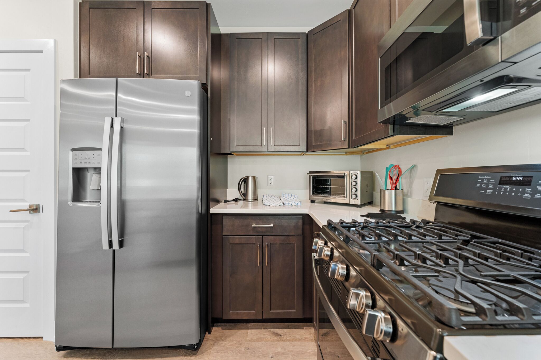 A kitchen with a stainless steel refrigerator, dark wooden cabinets, a gas stove, a microwave, and a kettle.