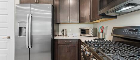 A kitchen with a stainless steel refrigerator, dark wooden cabinets, a gas stove, a microwave, and a kettle.