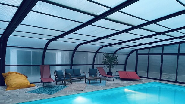 An indoor swimming pool with a glass ceiling, lounge chairs, and a yellow bean bag. - Villa Normande Piscine Intérieur et Jeux / Proche Honfleur et Deauville (Selles)
