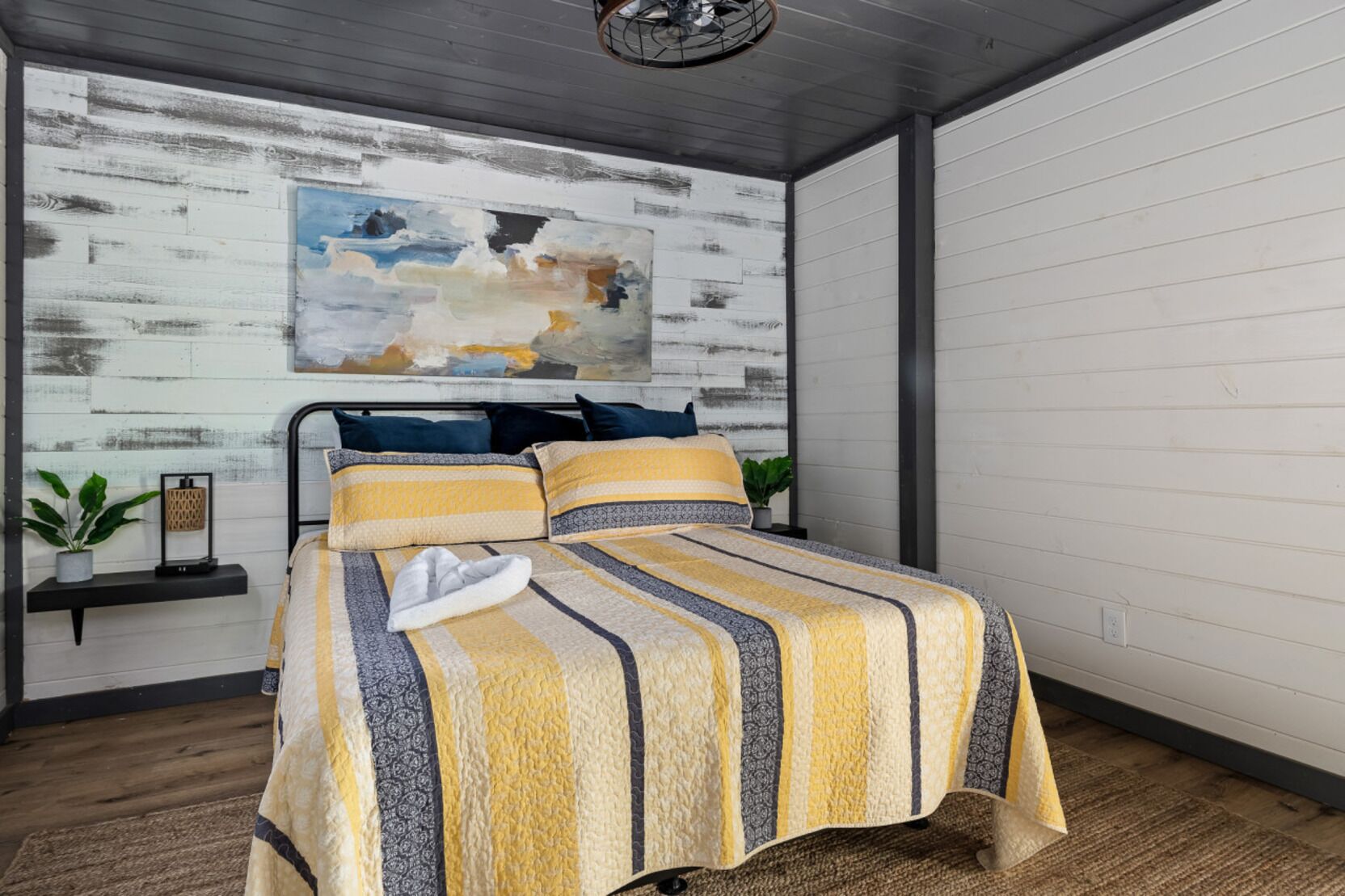 A bedroom with a bed, a painting, a shelf with plants, and a wooden floor.