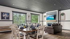 Un coin repas avec une table en bois dressée pour quatre personnes, entourée de chaises blanches. On y trouve également un canapé gris et un téléviseur à écran plat fixé au mur.