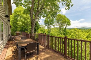 A wooden deck with a table and chairs, surrounded by a wooden railing, overlooking a lush green landscape. - Stags Leap at Yonahlossee (Boone)