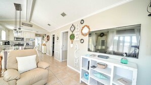 A spacious living room with a beige sofa, a TV on a white stand, and a kitchen in the background. - 264 Seahorse Lane - 3 Bedroom with Golf Cart Included! (Panama City Beach)