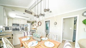 A dining area with a wooden table set for four, surrounded by white chairs. A kitchen with modern appliances is visible in the background. - 264 Seahorse Lane - 3 Bedroom with Golf Cart Included! (Panama City Beach)
