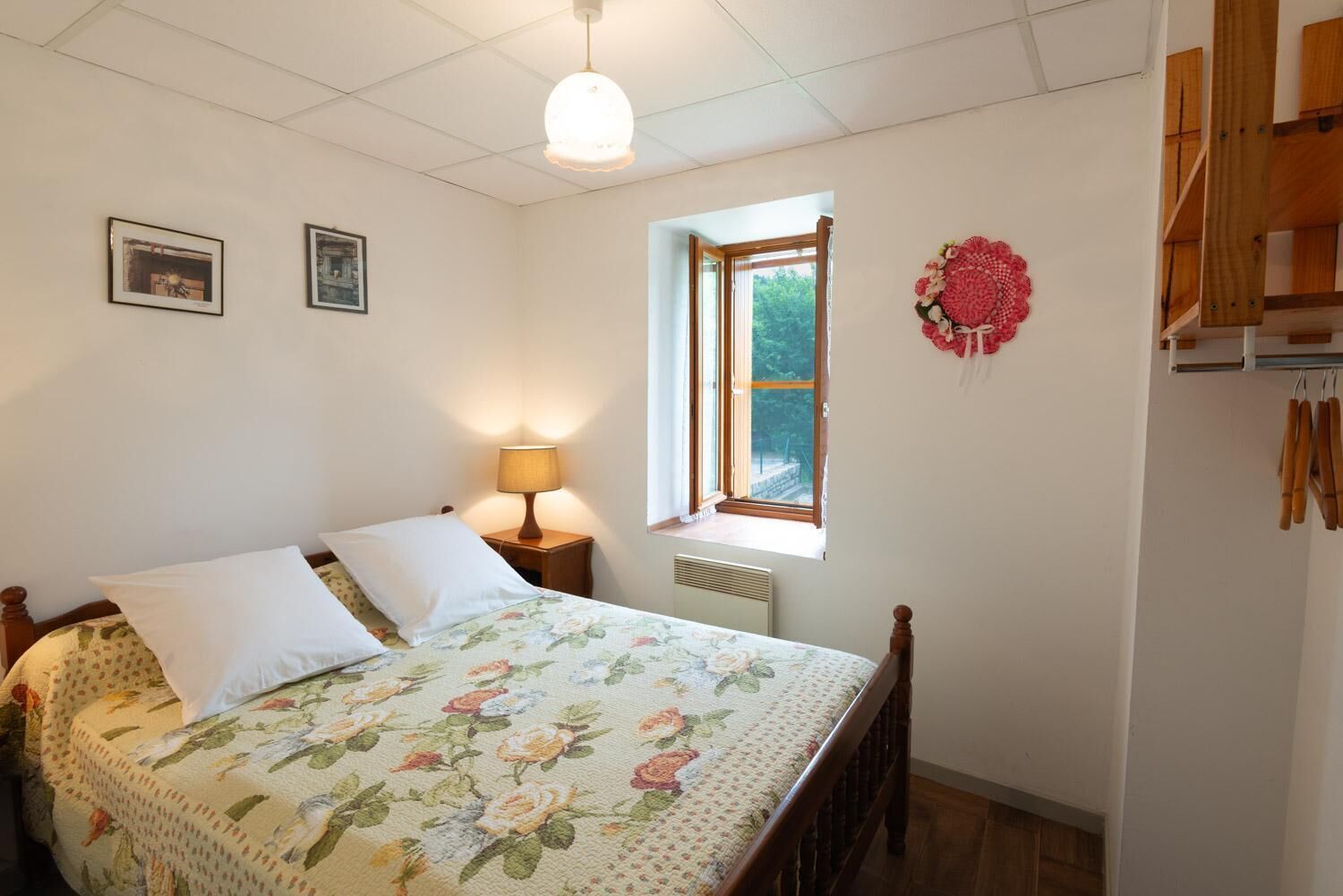 Un dormitorio con una cama, una mesita de noche con una lámpara, una ventana y un estante en la pared con utensilios de cocina.