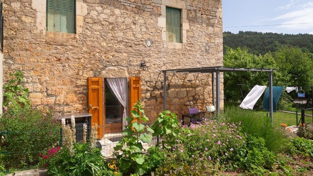 Un bâtiment en pierre avec un jardin à l’avant, un espace extérieur couvert et une vue sur une colline boisée.
