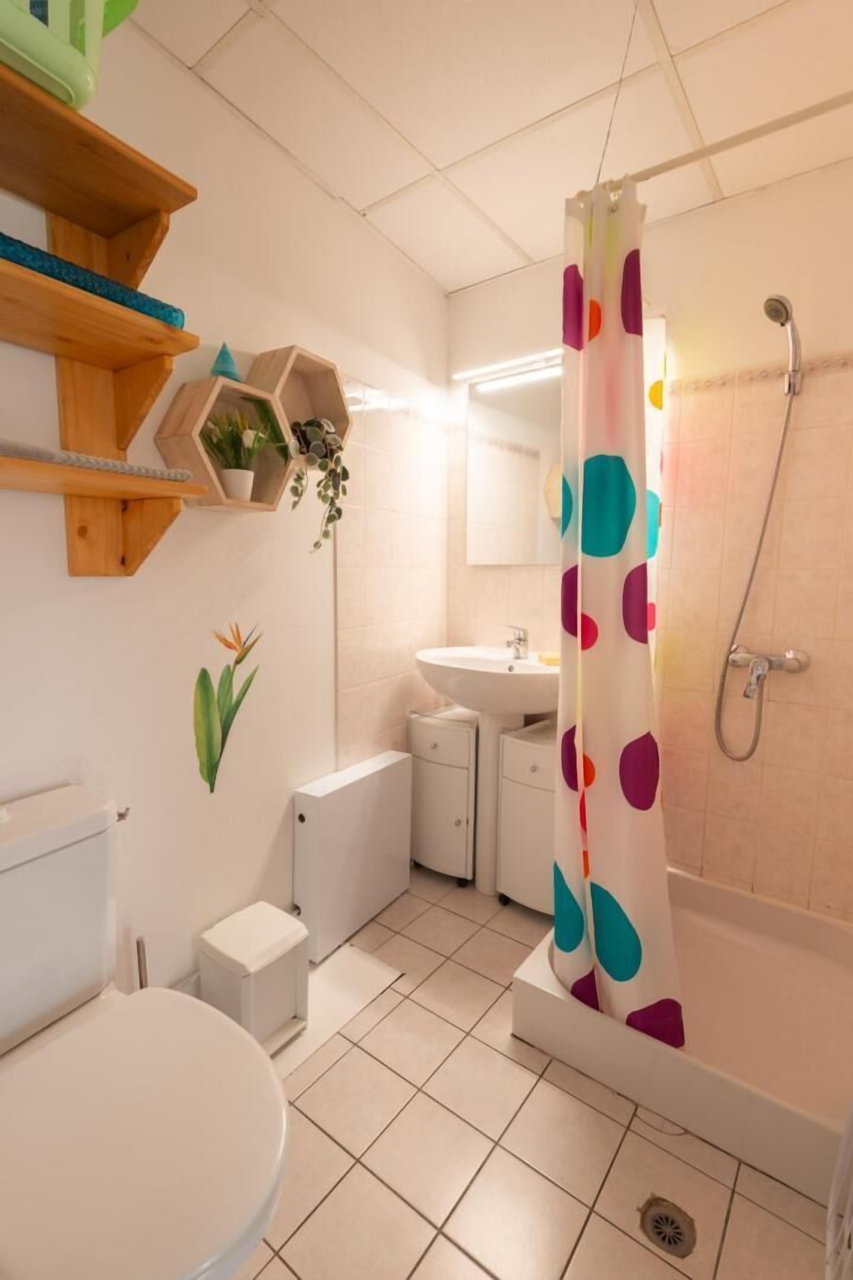 Une salle de bain avec des toilettes, un lavabo et une douche. On y trouve des étagères en bois ornées de plantes et un motif décoratif sur le mur.