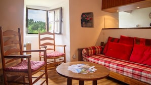 A cozy room with a plaid sofa, two wooden chairs, a round table with magazines, a window with a view of greenery, and a wall clock. - To the summits of the Gorges du Tarn (LAVAL DU TARN)
