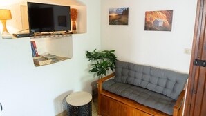 A cozy living room with a flat-screen TV, a cushioned bench, a small round table, and two framed paintings on the wall. - L'Oustal de Julie 3 people (LAVAL DU TARN)