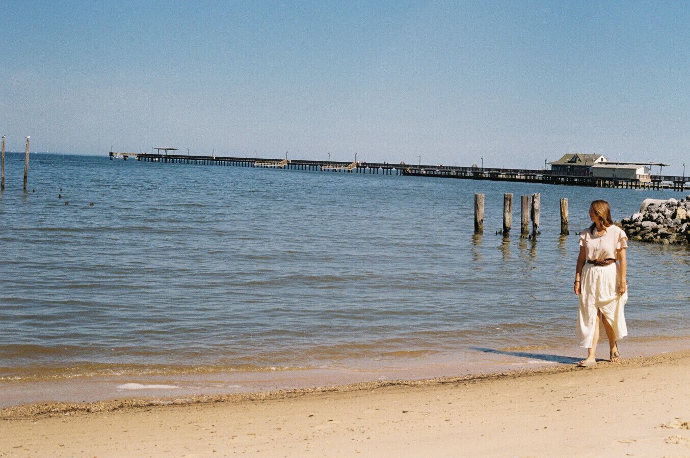 A beach with a pier extending into the water, a person walking along the shore, and a building in the background.