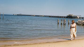 A beach with a pier extending into the water, a person walking along the shore, and a building in the background.