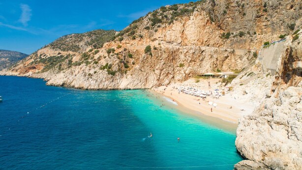 Ein Küstenstrand mit klarem türkisblauem Wasser, steinigen Klippen und einigen Sonnenschirmen im Sand.