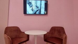 Ein Zimmer mit rosa Wänden, einem an der Wand montierten Flachbildfernseher und zwei braunen Stühlen um einem kleinen weißen Tisch.