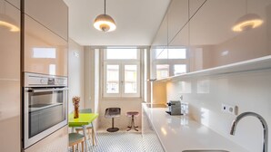 A modern kitchen with a built-in oven, a dining area with a green table and chairs, and a large window providing natural light. - VIVA Aliados Spacious & Bright Flat (Porto)