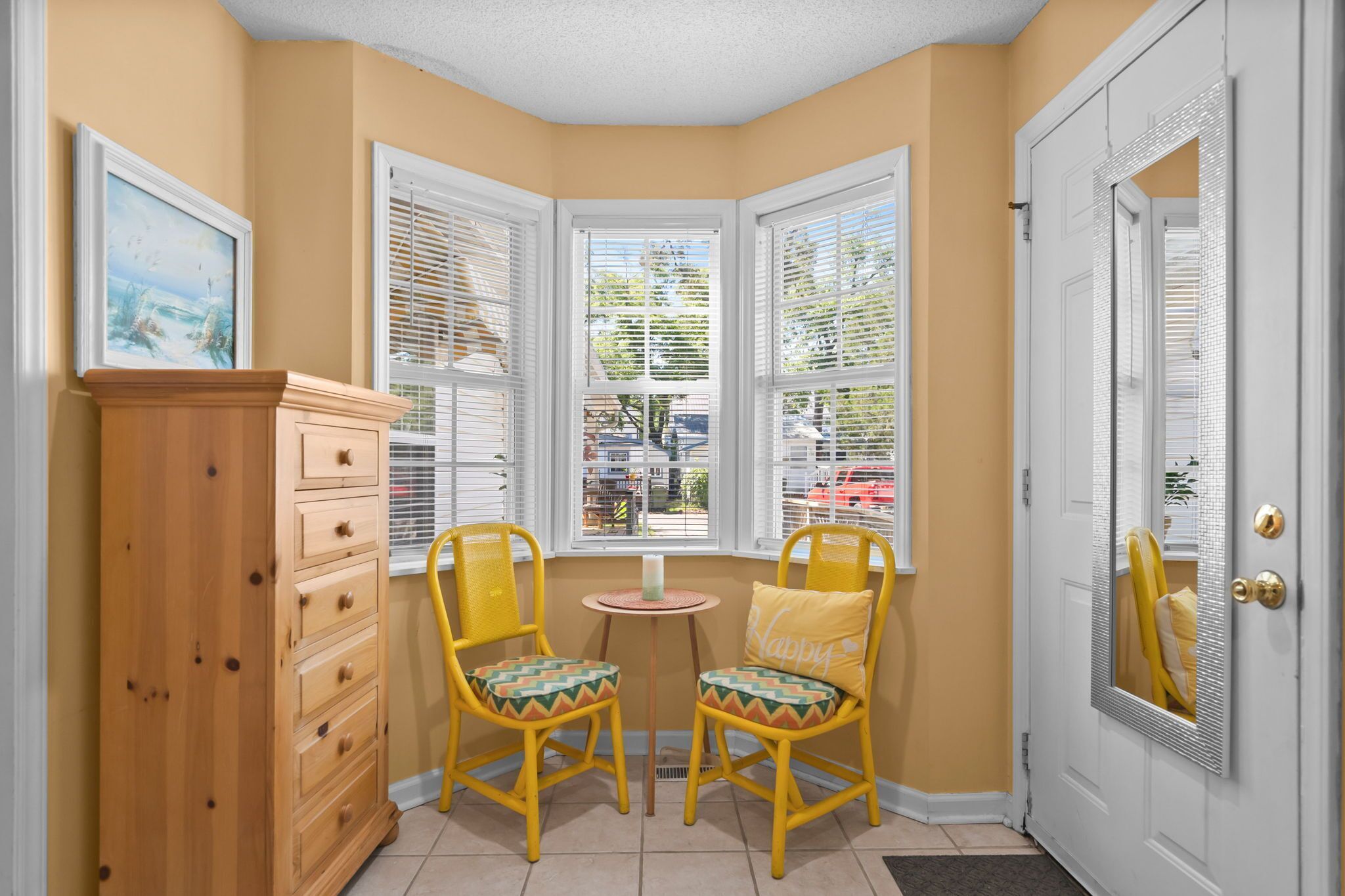 A corner room with a bay window, a wooden dresser, two yellow chairs, and a door with a mirror.